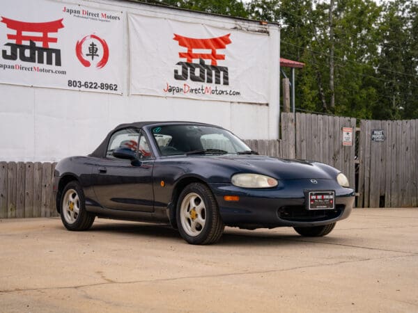 Blue convertible Mazda Miata parked in front of sign.